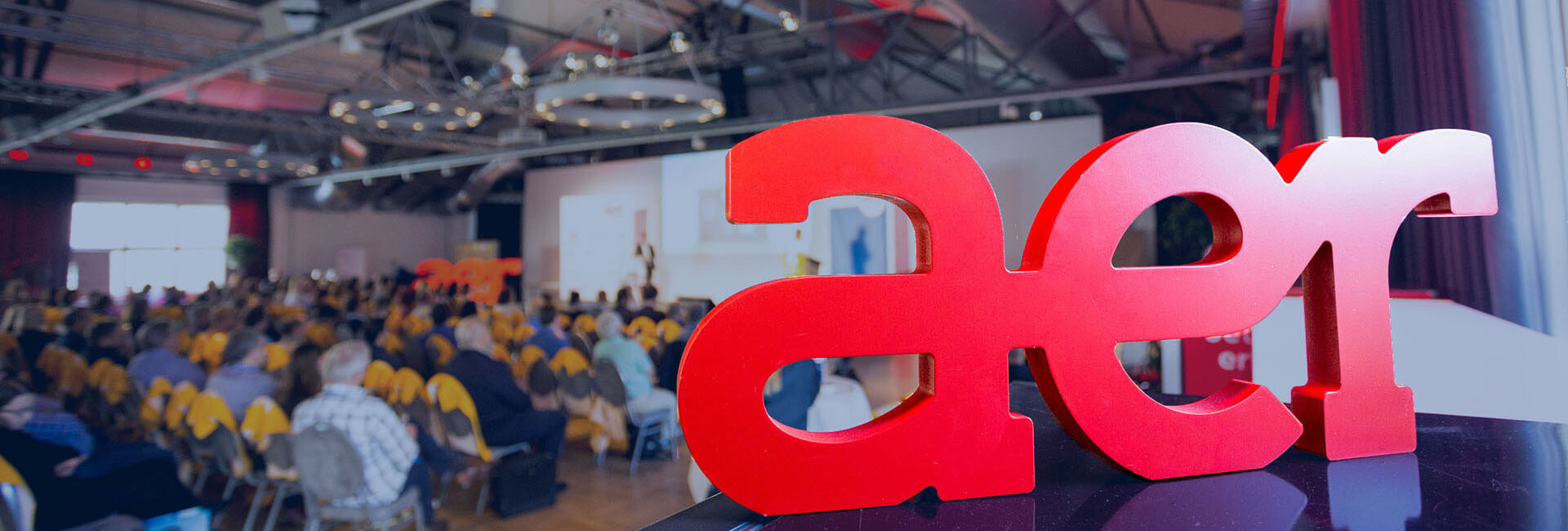 Red AER lettering on stage at an AER Kooperation event, with the audience in the background